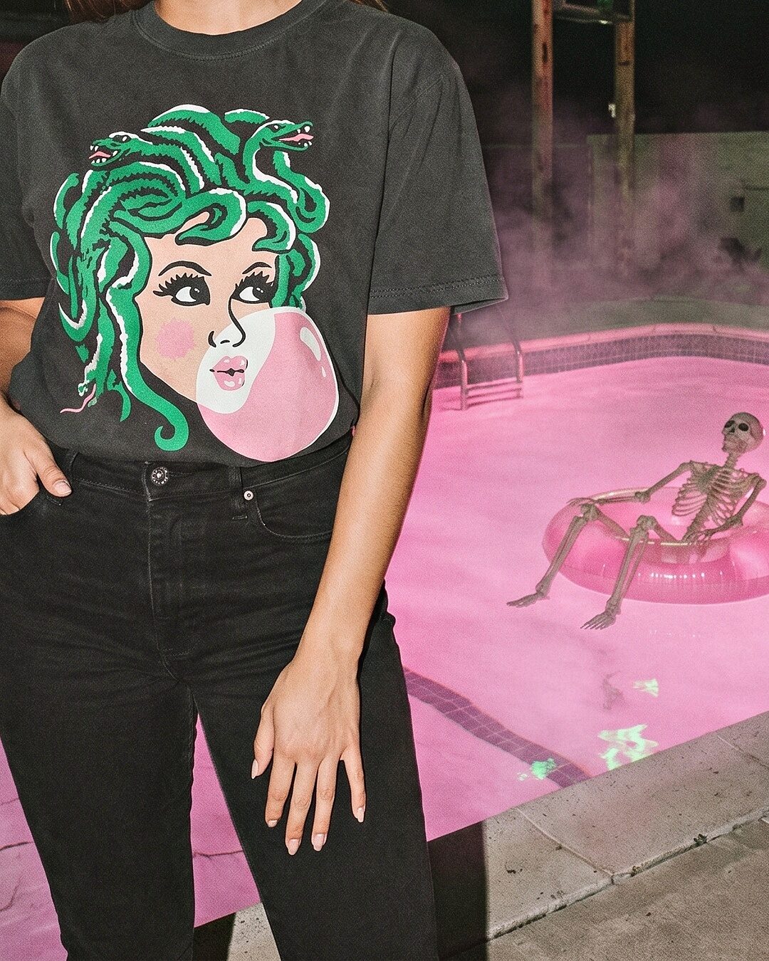 A woman in a Medusa Bubblegum graphic t-shirt standing beside a pink-lit pool with a skeleton floating on an inner tube.