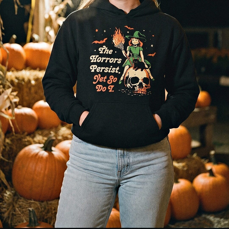 A woman in a black The Horrors Persist Yet So Do I hoodie standing in front of pumpkins and hay bales.