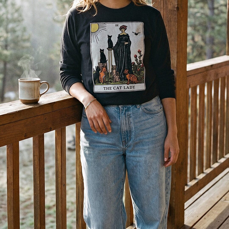 A woman in a black Cat Lady tarot card long sleeve t-shirt on a wooden porch with a mug of coffee.