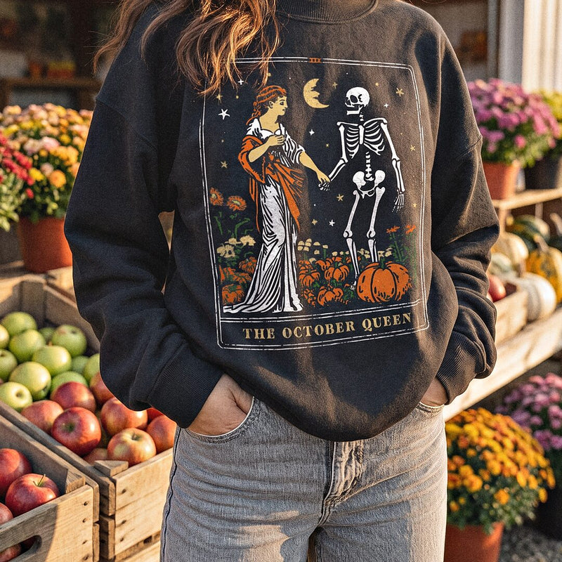 A woman in a black October Queen tarot card sweatshirt at a fall market with apples and mums.